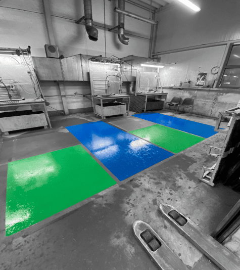 Farbige Bodenmarkierungen in Grün und Blau kennzeichnen Arbeitsbereiche in einer Industriehalle mit Maschinen.