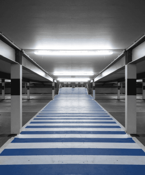 Weiße und blaue Streifen markieren einen breiten Fußgängerweg in einem beleuchteten Parkhaus.