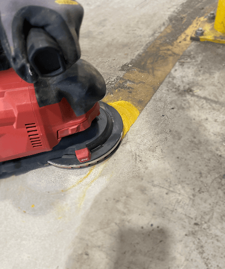 Bodenmarkierung in einer Halle, die mithilfe einer Bodenschleifmaschine entfernt wird. Dieser Prozess sorgt für die präzise Demarkierung von bestehenden Markierungen und bereitet den Boden für neue Markierungen vor