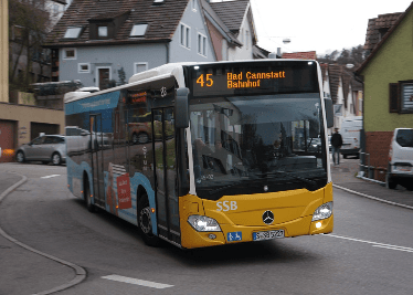 Linienbus fährt eine Kurve auf einer Straße, umgeben von urbaner Landschaft.
