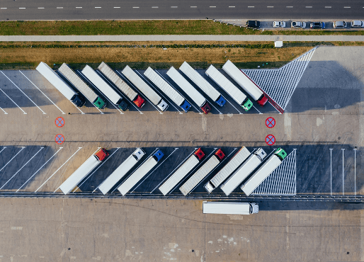 Luftaufnahme eines Parkplatzes mit zahlreichen schräg angeordneten LKW-Stellplätzen. Einige Bereiche sind durch Sperrflächen und Halteverbotsschilder markiert. Die LKWs stehen geordnet nebeneinander, während die verbotenen Zonen klar abgegrenzt sind.